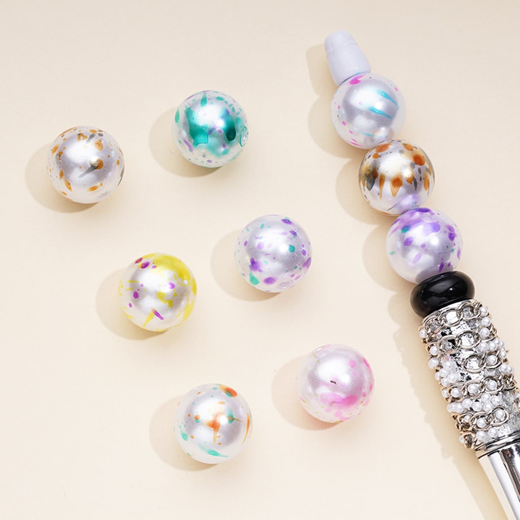 A collection of multicolored pearl design abstract splatter acrylic beads alongside a pen with a bead attached to its tip, displayed on a beige background.