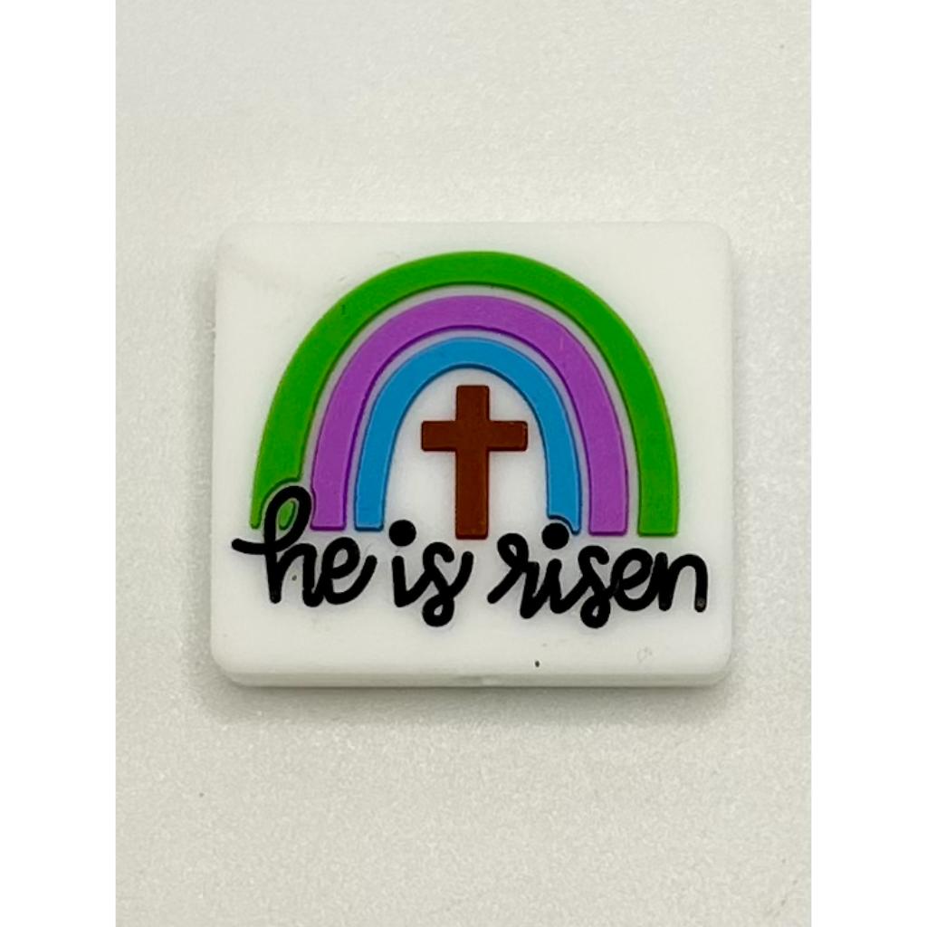 A silicone bead with a design that includes a rainbow, a cross, and the phrase 'He is risen' written in cursive.