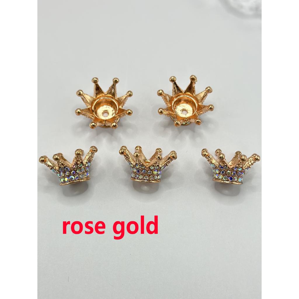 A collection of multicolored, crown-shaped bling bling spacers for beads displayed on a pink feather and musical score background.