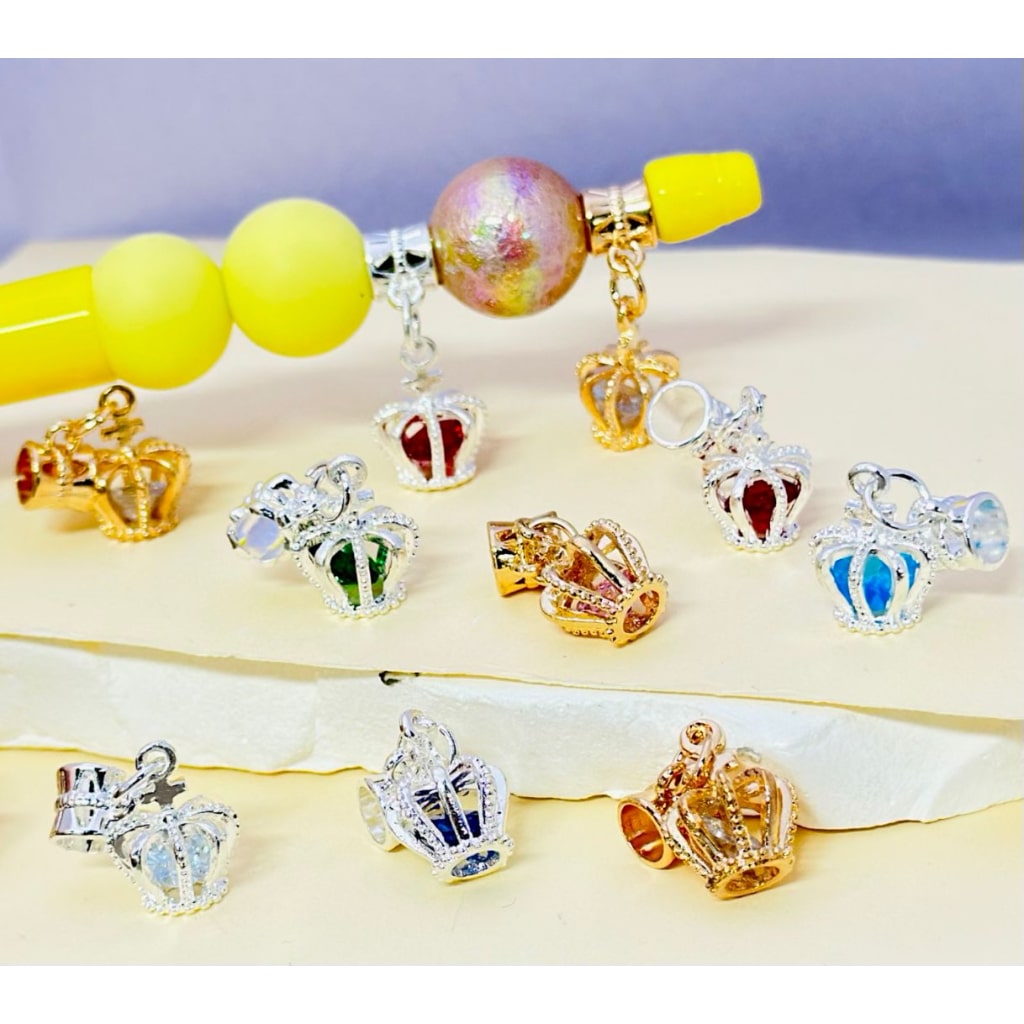 A variety of alloy crown-shaped beads with rhinestone pendants and jump rings attached to them, displayed on a surface.