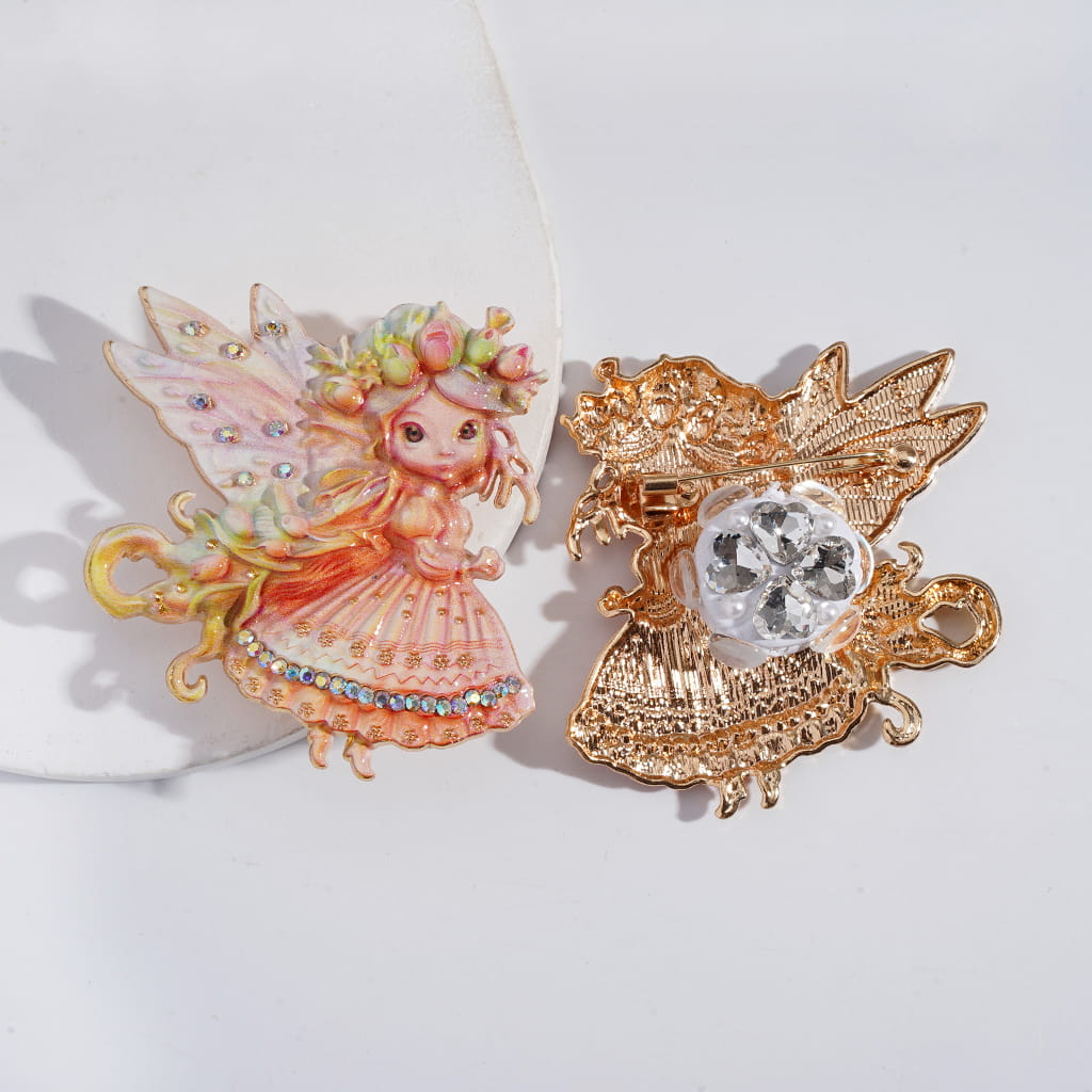 Two decorative pins, one fairy-like figure and one gold brooch with a gemstone, on a white background.