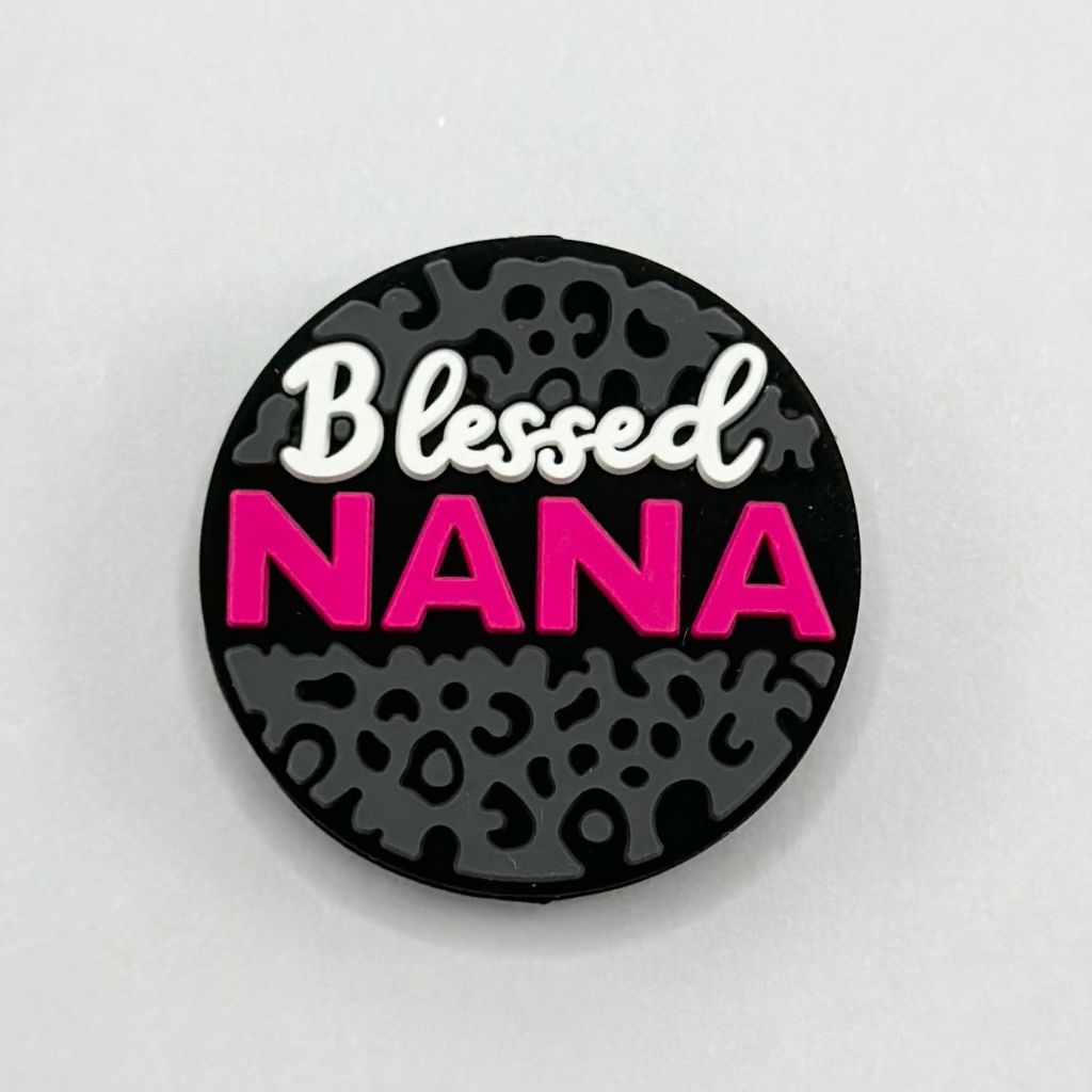 A round silicone bead with the text 'Blessed NANA' written on it, in white and pink colors against a black background with a pattern.