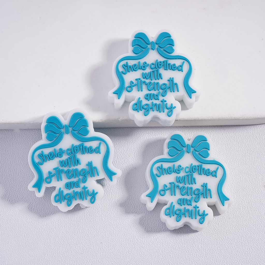 Three silicone beads with blue bows and the text 'She is clothed with strength and dignity' on a white surface.