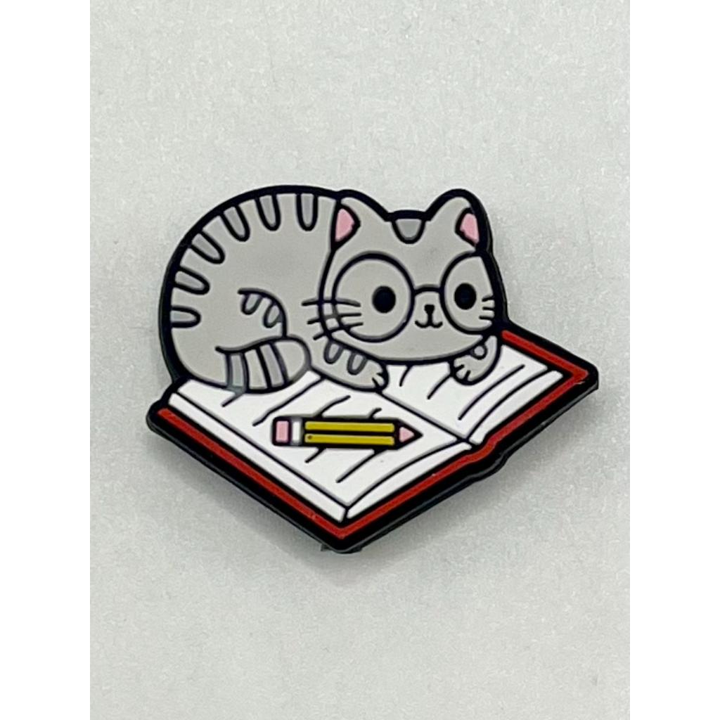 A silicone bead featuring a gray cat lying on an open book with a pencil placed on the book.