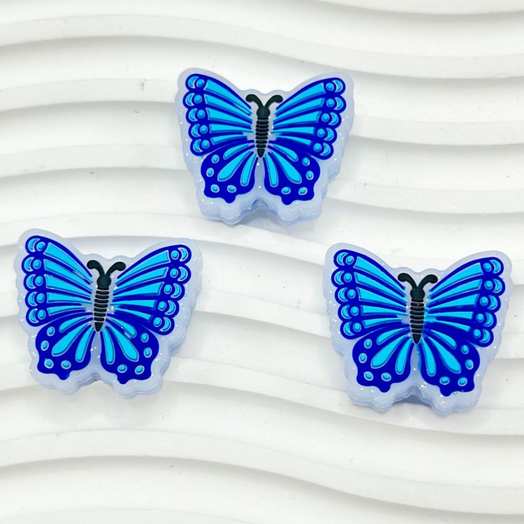 A set of three silicone beads in the shape of butterflies, with a dark blue body and blue patterned wings, arranged on a white surface.