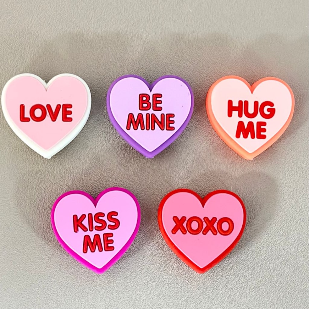 A collection of silicone beads in the shape of hearts with phrases related to love and affection written on them, including 'LOVE', 'BE MINE', 'HUG ME', 'KISS ME', and 'XOXO'. The beads are in shades of pink, red, white, and purple.