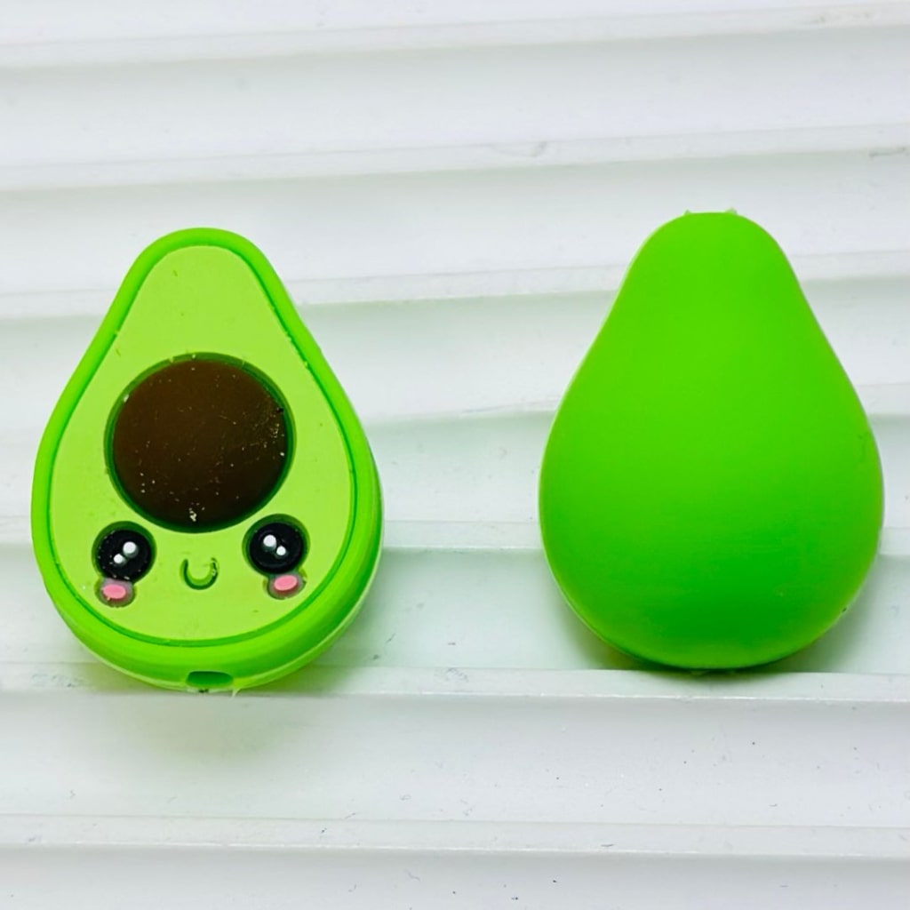 Two silicone beads shaped like avocados, one with a cute face design on the pit, on a white surface.