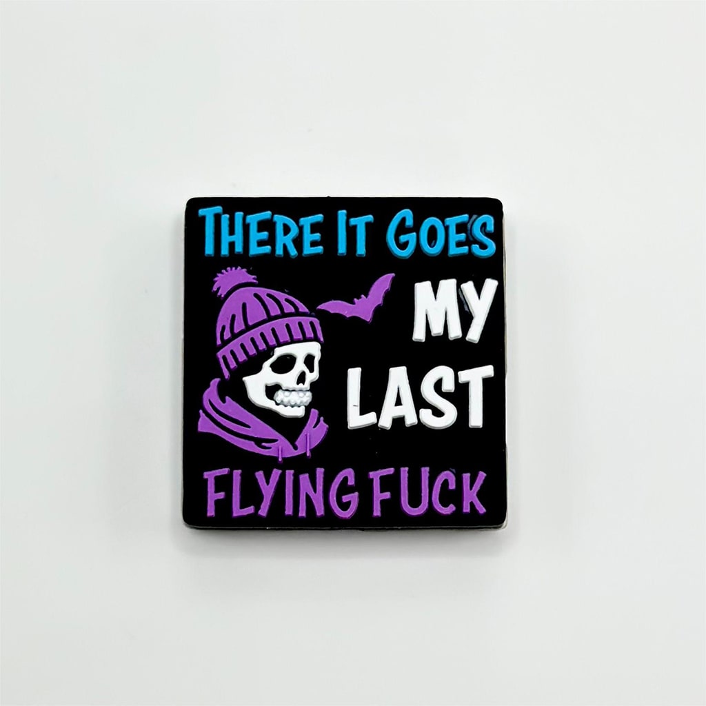 A black silicone focal bead with white and purple text that reads 'There It Goes My Last Flying Fuck' and an illustration of a skull wearing a hat.