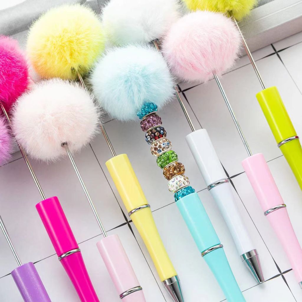 A collection of pens with colorful fluffy pompom toppers and elegant beadable designs on a metal rack.

