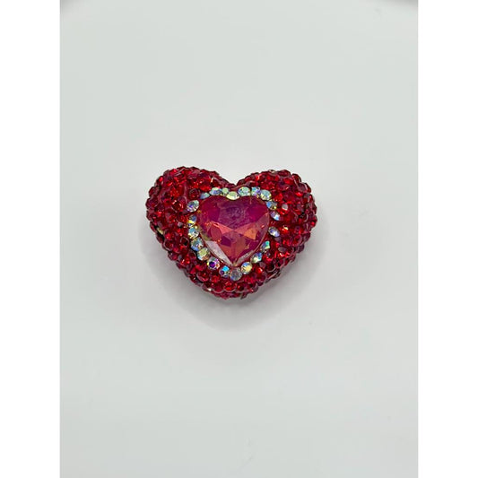 An assortment of heart-shaped clay beads in various colors, adorned with colorful rhinestones, displayed alongside a tool for crafting.

