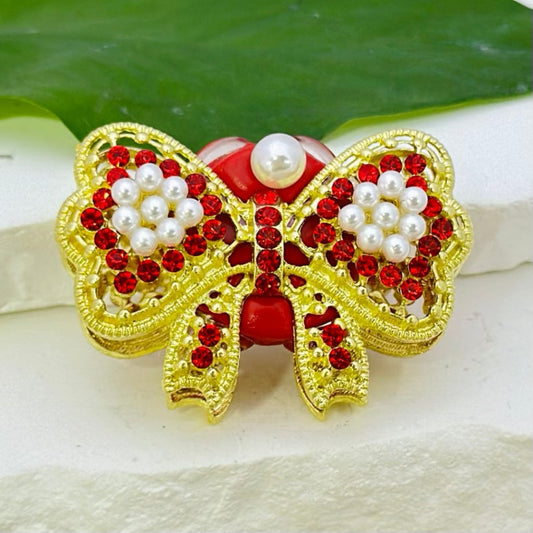A collection of gold alloy bowties, each adorned with colorful rhinestones, white pearls, and oval pearls, displayed against a leafy backdrop.

