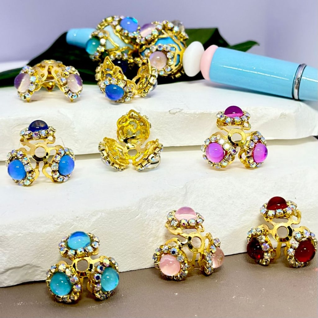 A collection of gold and silver bead caps with colorful rhinestones and large pearls designed for 16MM beads, displayed on a white surface.

