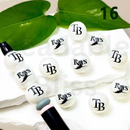 White beads with 'TB' and 'Rays' text on a white surface with green leaves in the background.

