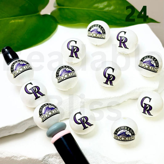 Colorful lip glosses with Colorado Rockies logo on a white surface with green leaves in the background.

