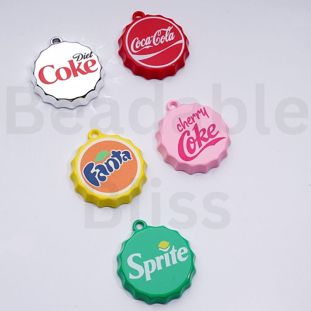 Five colorful bottle cap-shaped charms with Coca-Cola, Fanta, and Sprite logos on a white background.

