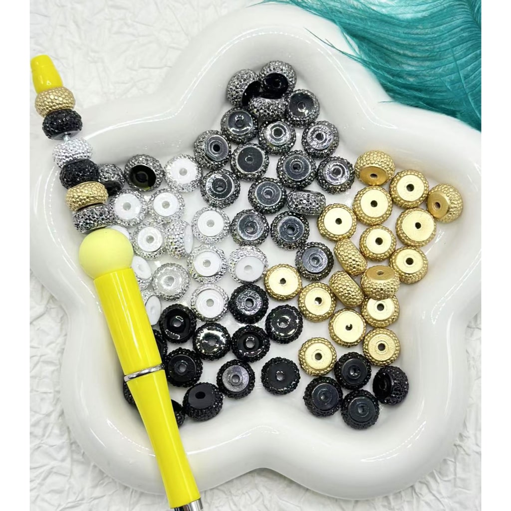 A collection of round rhinestone wheel spacers in silver, golden, and black colors displayed on a white surface with a yellow tool.

