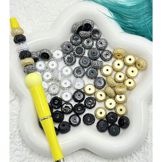 A collection of round rhinestone wheel spacers in silver, golden, and black colors displayed on a white surface with a yellow tool.

