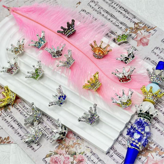 A collection of multicolored, crown-shaped bling bling spacers for beads displayed on a pink feather and musical score background.

