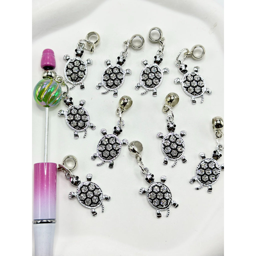 A collection of silver-colored turtle-shaped alloy charms with clear rhinestones displayed alongside a pen for scale.


