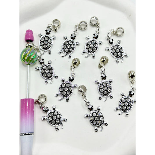 A collection of silver-colored turtle-shaped alloy charms with clear rhinestones displayed alongside a pen for scale.

