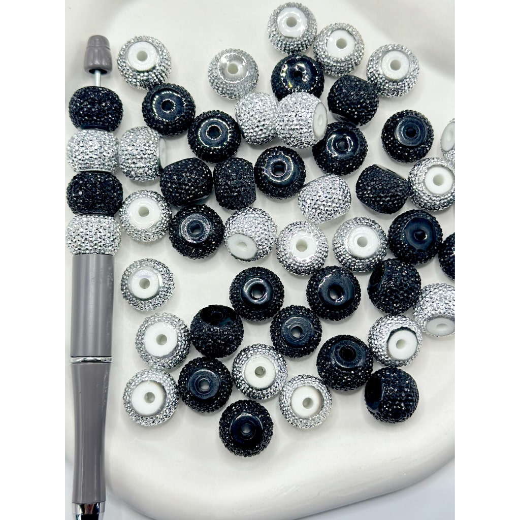 A collection of round acrylic beads in silver and black, with a sparkling rhinestone wheel design, displayed on a white surface alongside a tool for crafting.

