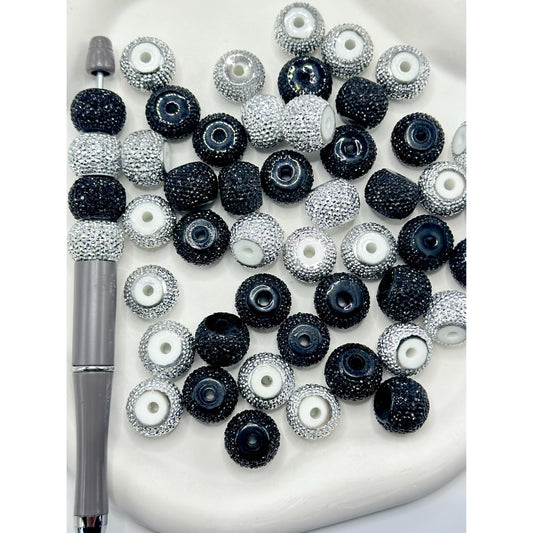 A collection of round acrylic beads in silver and black, with a sparkling rhinestone wheel design, displayed on a white surface alongside a tool for crafting.

