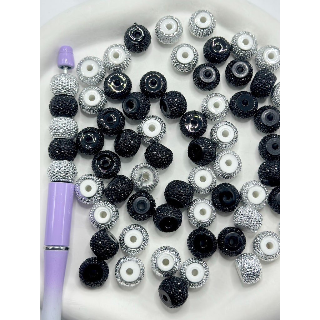 A collection of round acrylic beads in black, silver, and white colors with visible rhinestone embellishments, displayed alongside a purple stylus on a white surface.

