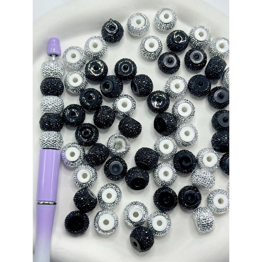 A collection of round acrylic beads in black, silver, and white colors with visible rhinestone embellishments, displayed alongside a purple stylus on a white surface.


