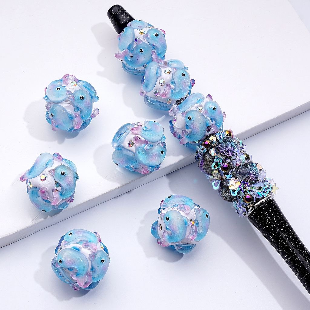 A collection of blue dolphin-shaped clay beads with AB rhinestones, approximately 20mm in size, displayed alongside a beadable pen.

