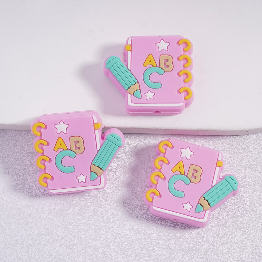 Three pink book-shaped clips with ABC letters and colorful pencils on a light purple background