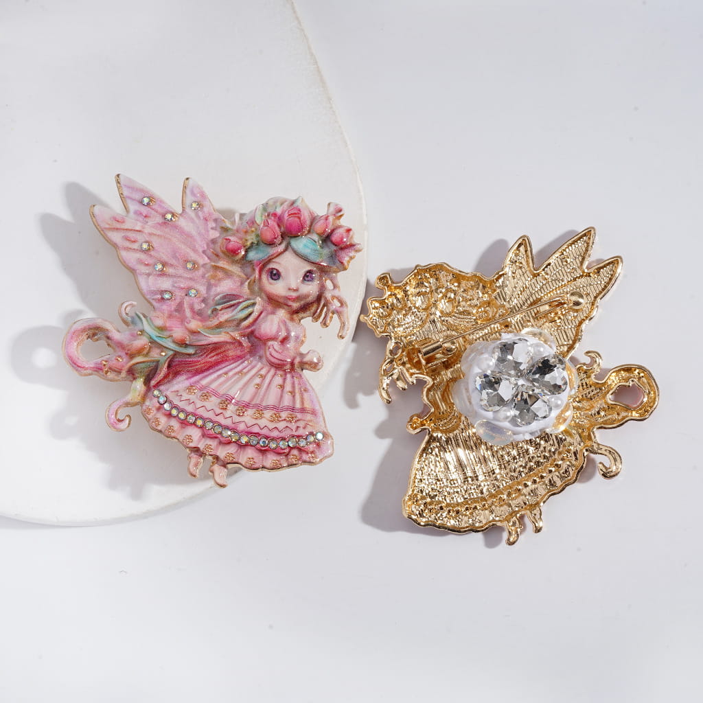 Two decorative pins on a white background, one pink fairy and one gold brooch with a central gemstone.