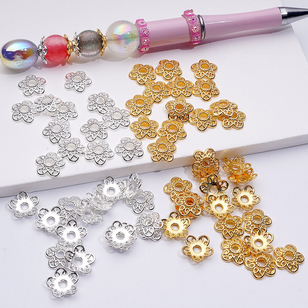 Gold, silver, and rose gold flower-shaped beads on a white surface with a pen.

