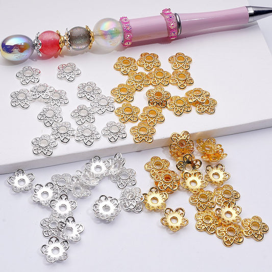 Gold, silver, and rose gold flower-shaped beads on a white surface with a pen.


