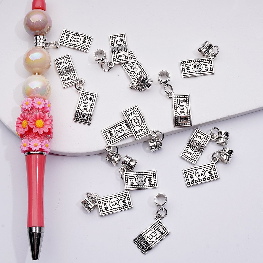 Silver money bill charms scattered on a white surface with a pink pen and beads.

