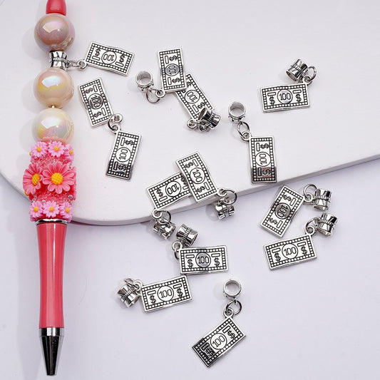 Silver money bill charms scattered on a white surface with a pink pen and beads.

