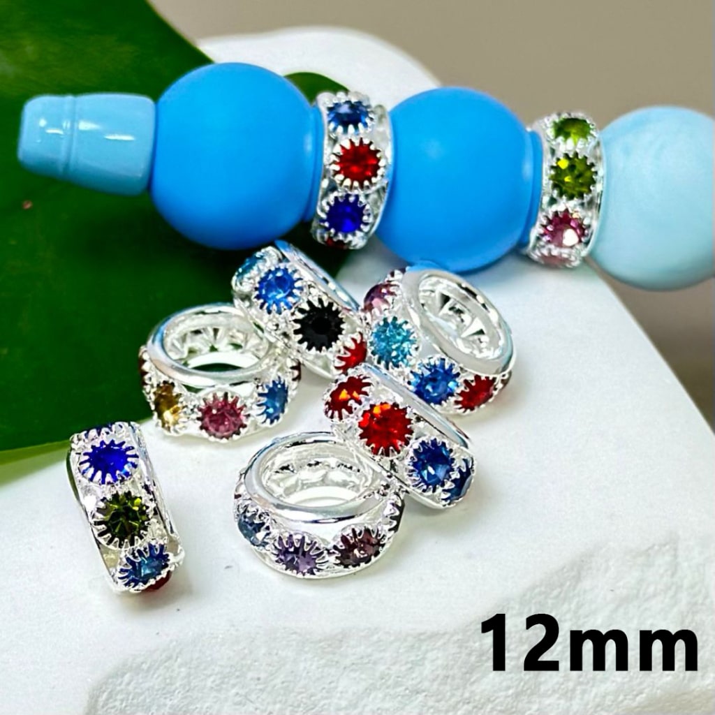 Silver alloy ring spacers with colorful mini rhinestones embedded in them, displayed alongside a blue bead and a green leaf for scale reference, indicating the size of 12mm.

