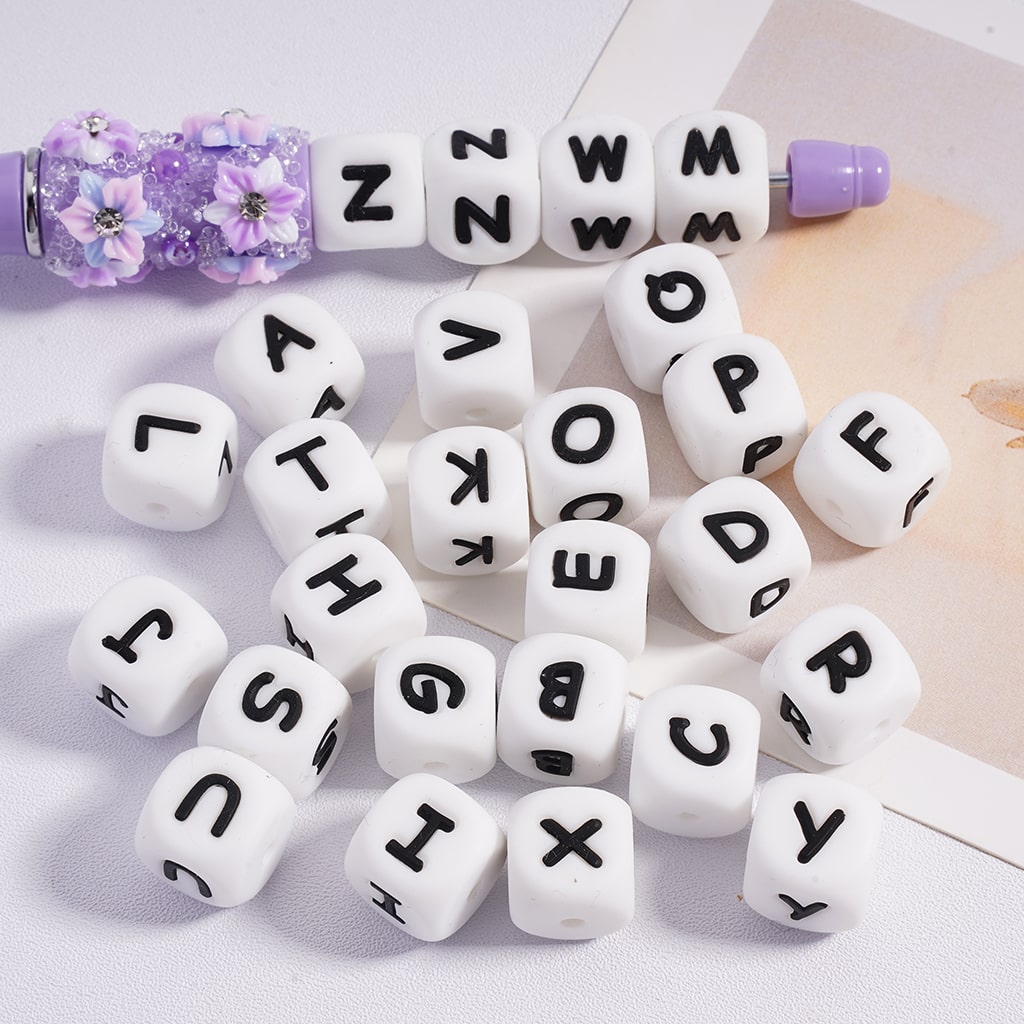 A collection of white silicone letter beads laid out on a surface, with each bead featuring a black alphabet letter.

