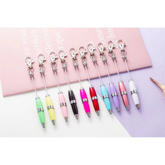 A collection of metal keychain pens with bead decorations, featuring a lobster clasp, displayed on a pink surface.

