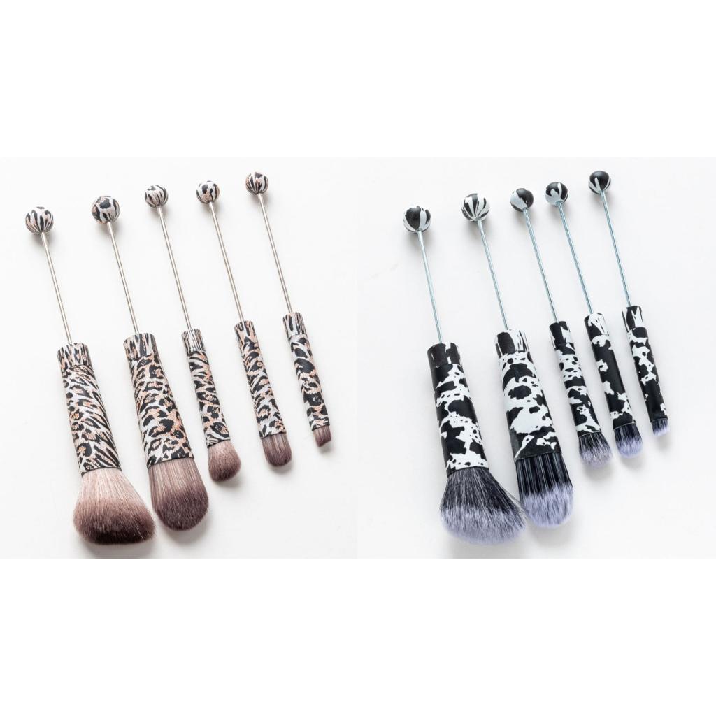 A collection of makeup brushes with animal print handles, including leopard and jaguar designs, displayed in two rows with brushes with silver bead embellishments on the handles.

