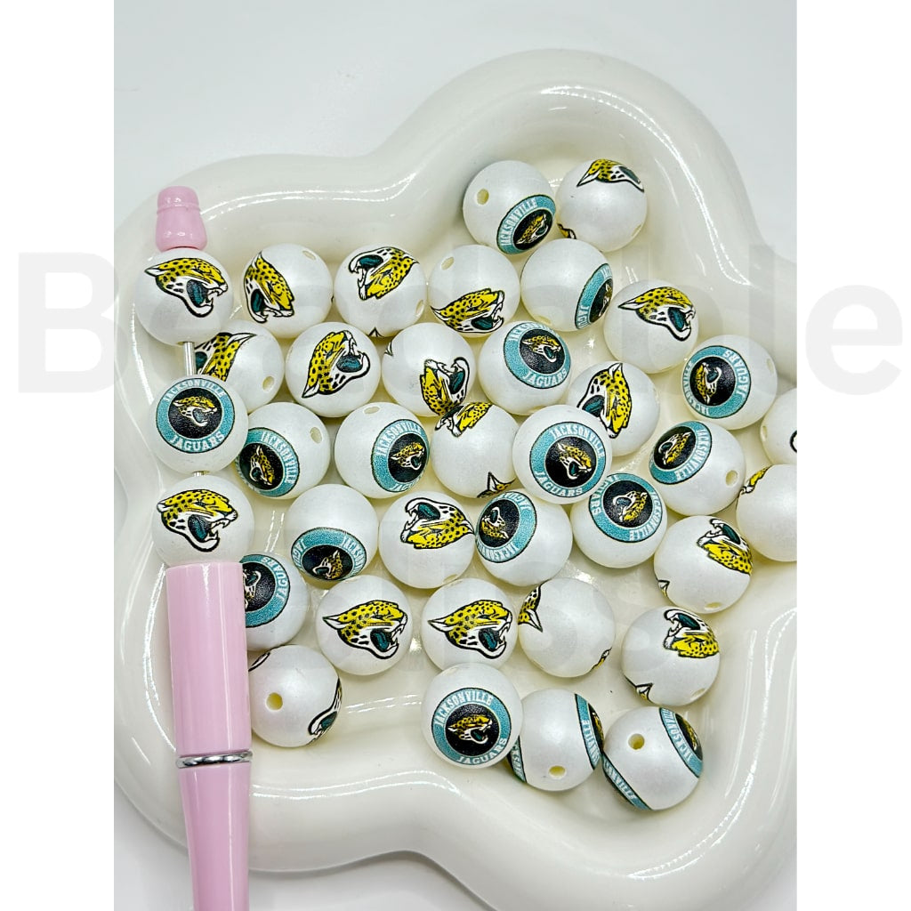 White beads with sports team logos on a white heart-shaped dish with a pink pen.

