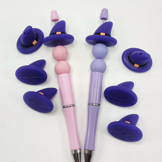 A collection of purple witch hat shaped silicone beads in various sizes, displayed alongside two pens with similar hat designs on them.

