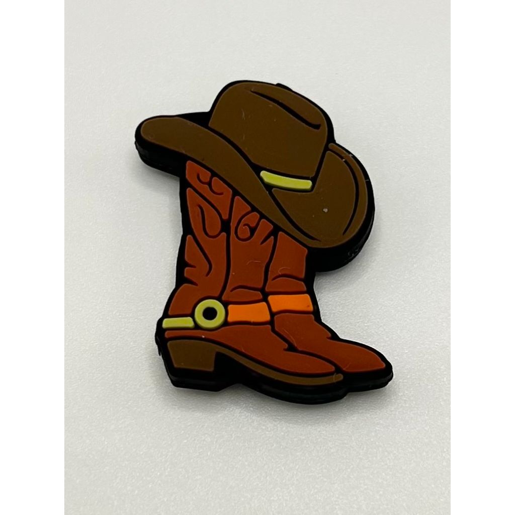 A silicone bead in the shape of a cowboy hat placed on top of a pair of boots, primarily brown in color with multistorey beadable pens.


