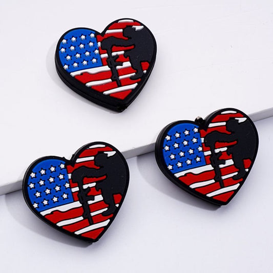 The image features a collection of silicone beads, each shaped like a heart with the American flag design. The beads are laid out on a white surface and appear to be used for crafting or jewelry making.

