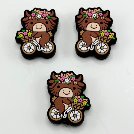 A set of silicone beads featuring a brown cow riding a bike with small flowers.

