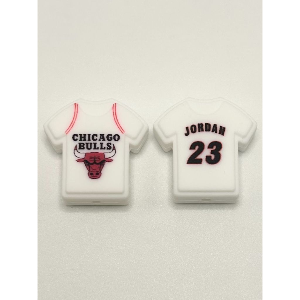 Two silicone beads designed to look like white Chicago Bulls basketball jerseys with the number 23 and the name 'Jordan' printed on the back, one with a bull's head logo.

