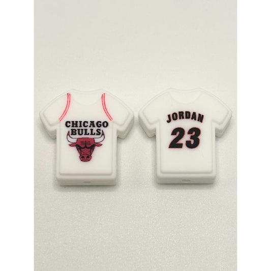 Two silicone beads designed to look like white Chicago Bulls basketball jerseys with the number 23 and the name 'Jordan' printed on the back, one with a bull's head logo.

