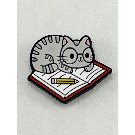 A silicone bead featuring a gray cat lying on an open book with a pencil placed on the book.

