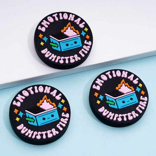 The image shows three round silicone beads with a design that resembles a cartoonish dumpster on fire, and the text 'EMOTIONAL DUMPSTER FIRE'. The background is black, and the beads are displayed on a light blue surface.

