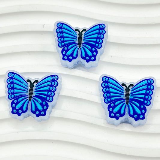A set of three silicone beads in the shape of butterflies, with a dark blue body and blue patterned wings, arranged on a white surface.


