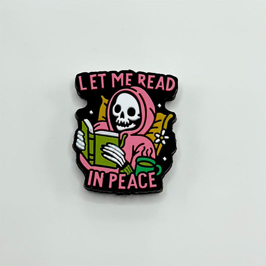 A silicone bead featuring a skeleton skull with the phrase 'LET ME READ IN PEACE' written on it, primarily in black with pink and green accents.

